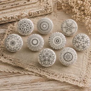 May include: Nine white ceramic cabinet knobs with grey decorative patterns. The knobs feature various designs, including floral, geometric, and mandala-style motifs. They are arranged on a beige lace-trimmed cloth, suggesting a home decor theme.