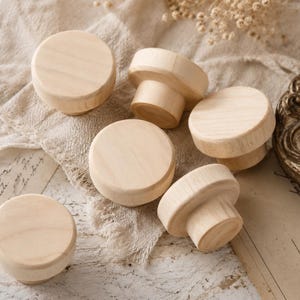 May include: A collection of unfinished wooden knobs in various shapes and sizes. The knobs are light-colored and appear to be made of natural wood. They are arranged on a beige, textured fabric, suggesting a craft or DIY project.