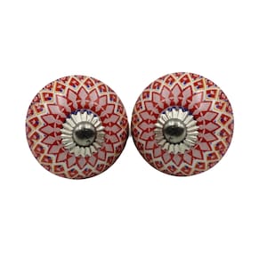 May include: Two decorative cabinet knobs. Each knob features a round, ceramic design with a red and white floral pattern. The knobs have a silver-colored metal base and are set against a white background.