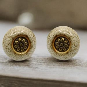 Puede incluir: Dos pomos de armario redondos, moteados, de color crema, con un diseño floral de color latón en el centro. Los pomos se colocan sobre un fondo neutro, mostrando sus detalles decorativos.