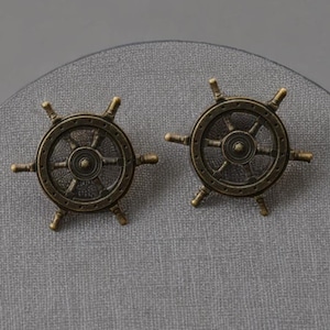 May include: Two antique brass-colored ship wheel cabinet knobs. Each knob features a detailed wheel design with spokes and a central hub. The knobs are set against a textured gray background, evoking a nautical theme.