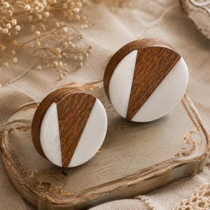 May include: Two decorative wooden cabinet knobs with a geometric design. Each knob features a half-circle of light-colored wood and a white, triangular inlay. The knobs are displayed on a light-colored wooden tray.
