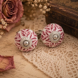 Può includere: Due pomelli rotondi in ceramica per mobili con base bianca e un motivo a foglie ripetuto in rosa scuro. Ogni pomello presenta un rosone centrale in metallo color argento con un piccolo perno circolare al centro come elemento decorativo.