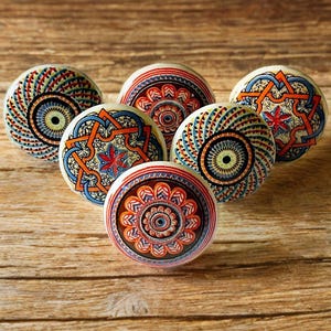 Könnte beinhalten: Sechs Keramik-Knöpfe mit farbenfrohen, komplizierten Mandala-Designs. Die Knöpfe sind rund und haben einen weißen Hintergrund mit orangefarbenen, blauen und roten Akzenten.