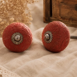 May include: Two red, textured cabinet knobs with silver-colored decorative centers. The knobs are round and have a rustic appearance. They are set against a neutral-toned fabric background, suggesting a home decor theme.