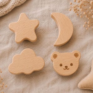 以下が含まれることがあります： 星、三日月、雲、クマの形をした木製のおもちゃのセット。おもちゃは明るい色の木材で作られており、ニュートラルな色の布の上に配置されています。クマには彫られた顔の特徴があります。