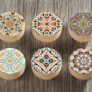 May include: Six small, round containers with decorative lids. The lids feature intricate, colourful mandala patterns in shades of orange, blue, green, and cream. The containers have a light beige base, set against a wood grain background.