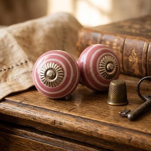Puede incluir: Dos pomos de cerámica a rayas rosas y blancas con detalles metálicos plateados. Una dedal y llaves antiguas descansan sobre una superficie de madera junto a un libro antiguo y un paño de lino beige.