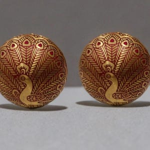 Może przedstawiać: Dwie złote, kuliste kolczyki z motywem pawia. Kolczyki mają czerwono-złoty wzór, ze szczegółami ciała i piór ogona pawia. Kolczyki są okrągłe i mają gładkie, wypolerowane wykończenie.