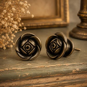 May include: Two antique bronze cabinet knobs with a rose design. The knobs are round with detailed petal patterns and a central square design. One knob has a threaded post for installation. Decorative hardware.