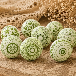 May include: Eight white ceramic cabinet knobs with green decorative patterns. The knobs are arranged on a distressed wooden surface, featuring floral and geometric designs. Each knob is approximately 4 cm in diameter.