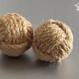 Pode incluir: Duas bolas decorativas de corda em estilo n&aacute;utico. As bolas s&atilde;o feitas de corda de cor natural, fortemente enrolada e amarrada em forma redonda. A textura da corda &eacute; vis&iacute;vel. Est&atilde;o sobre uma superf&iacute;cie cinza.
