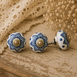 May include: Three decorative ceramic cabinet knobs. The knobs are white with a blue floral design and a gold-coloured centre. The knobs are on a wooden surface.