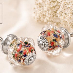 Peut inclure: Deux boutons de meuble en verre transparent avec des éclats colorés rouges, bleus et dorés. Les boutons ont une base et une vis en métal argenté. Le texte "O & K" est dans un encadré blanc en haut à gauche. Quincaillerie décorative.