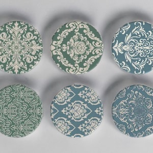 May include: Six circular coasters featuring white damask patterns on teal and green backgrounds. The coasters are arranged in two rows of three, displaying different designs. They are likely made of ceramic or a similar material, perfect for protecting surfaces.