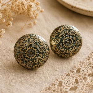 May include: Two round, decorative cabinet knobs with a dark teal background and gold floral design. The knobs are placed on a beige fabric surface, with a lace trim and dried flowers in the background.