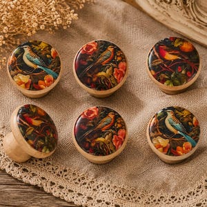 Puede incluir: Seis pomos de armario de madera con coloridos diseños de pájaros y flores. Los pomos tienen un acabado brillante y están colocados sobre un fondo de tela neutra. Los pájaros tienen plumaje azul, naranja y amarillo, y las flores son naranjas y rojas.