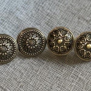Puede incluir: Cuatro botones antiguos de latón con diseños florales y de volutas detallados. Dos botones presentan un patrón de girasol, mientras que los otros dos tienen un motivo adornado y arremolinado. Los botones son redondos y tienen un aspecto ligeramente envejecido.