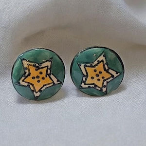 Pode incluir: Um par de brincos de estrela verde e amarelo. As estrelas têm um contorno branco e pontos pretos.