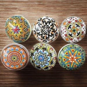 Puede incluir: Seis pomos de cerámica decorativos con patrones coloridos e intrincados. Los pomos presentan una variedad de diseños, incluidos motivos florales y geométricos, en tonos de azul, naranja, amarillo y negro. Están dispuestos sobre una superficie de madera.