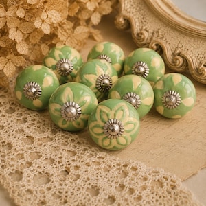 Puede incluir: Un conjunto de diez pomos de cerámica verdes y crema. Cada pomo presenta un diseño floral y una base de metal plateado. Los pomos están dispuestos sobre una superficie crema con un borde de encaje. Cada pomo mide unos 4 cm de diámetro.