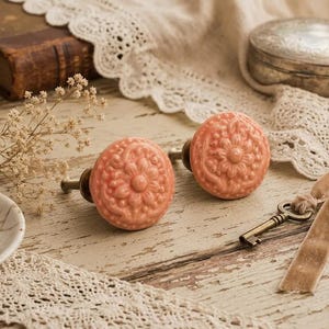 Puede incluir: Dos pomos de cerámica color coral con diseño floral. Se encuentran sobre una superficie de madera rústica y desgastada, junto a una llave antigua y adornos de encaje. También se ven un plato pequeño con pétalos de rosa secos y una botella pequeña.