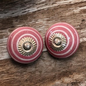 Puede incluir: Dos pomos de cerámica a rayas rosas y blancas con detalles metálicos dorados. Los pomos tienen un diseño circular con rayas concéntricas y un centro decorativo. Sobre un fondo de madera.