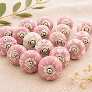 May include: A selection of pink and white ceramic cabinet knobs with floral and patterned designs. Each knob has a silver-coloured metal base. The knobs are arranged on a light-coloured fabric surface.