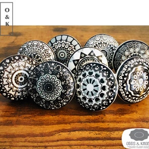 Op de afbeelding: Een set van acht zwarte en witte keramische kastknoppen met ingewikkelde mandala-ontwerpen. De knoppen zijn in een cirkel op een houten oppervlak gerangschikt.