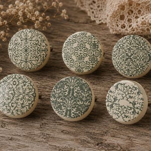 May include: Six cream-coloured cabinet knobs featuring a dark green, ornate pattern. The knobs are displayed on a rustic wooden surface, highlighting their intricate designs. Each knob is designed for use in home decor.
