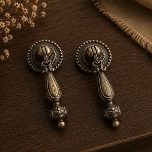 May include: Two antique brass drawer pulls with ornate detailing. Each pull features a circular backplate with a beaded edge, a decorative handle, and a detailed base. The pulls are set against a dark wood surface.
