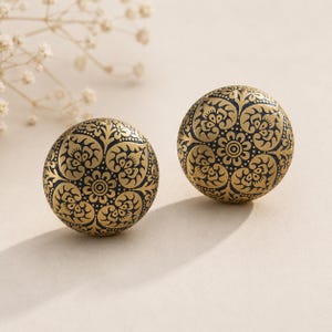 May include: Two round, gold-colored decorative knobs with intricate black floral patterns. The knobs are spherical and feature a detailed design of swirling leaves and flowers. The background is a soft, neutral color, highlighting the ornate details of the knobs.