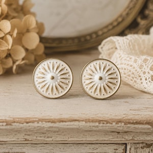 Könnte beinhalten: Zwei dekorative Schrankknöpfe in Weiß und Gold mit Sonnenstrahlen-Design. Die runden Knöpfe haben ein strukturiertes, erhabenes Muster und einen goldfarbenen Rand. Sie liegen auf einer rustikalen, hellen Holzoberfläche.