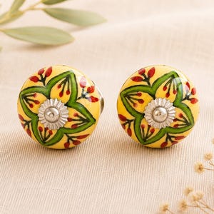 May include: Two yellow cabinet knobs with a floral design. The knobs have a green and red floral pattern and a silver metal centre. The knobs are on a cream-coloured surface.
