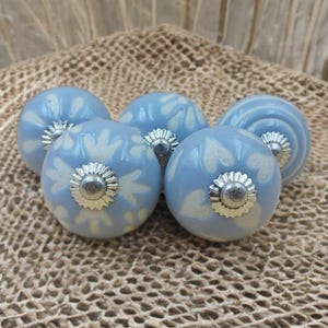 May include: Five pale blue ceramic cabinet knobs with white floral patterns and silver metal fixings. The knobs are spherical and displayed on a textured, neutral-toned surface.