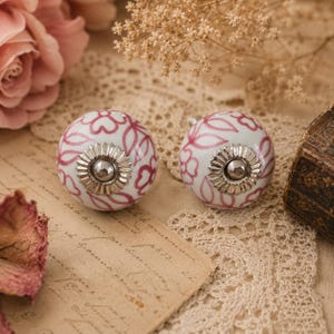 May include: Two white ceramic cabinet knobs with a pink floral pattern and silver hardware. The round knobs feature a decorative design. These knobs are likely for a drawer or cabinet.