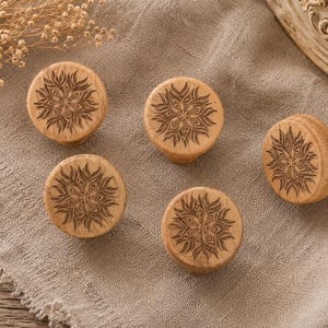 Peut inclure: Cinq boutons de meubles en bois clair avec un motif floral gravé sur le dessus. Les boutons sont ronds avec une base cylindrique courte. Ils sont disposés sur une surface en lin beige.