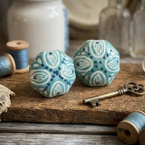 May include: Two turquoise ceramic cabinet knobs with white and navy blue floral designs. A vintage brass key and spools of thread are also on the wooden surface. Decorative hardware for furniture.