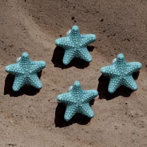Könnte beinhalten: Vier türkisfarbene, sternförmige Schrankknöpfe mit strukturierter Oberfläche. Die Knöpfe sind auf einer sandigen Oberfläche angeordnet und erinnern an ein Küsten- oder maritimes Thema. Jeder Knopf hat einen kleinen, weißen Montagepfosten.