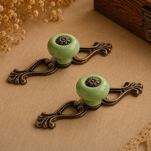May include: Two antique-style cabinet pulls. Each pull features a light green ceramic knob with a bronze-coloured floral design and a bronze-coloured ornate backplate. The pulls are displayed on a neutral-coloured surface.
