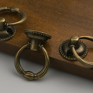 Puede incluir: Tres tiradores de cajón antiguos de bronce con tiradores de anillo. Los tiradores tienen un diseño decorativo y están colocados sobre una superficie de madera. Los herrajes son probablemente para armarios o muebles.