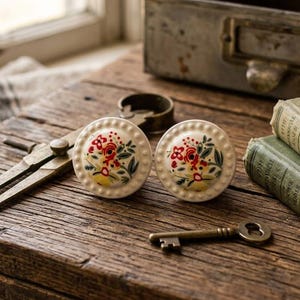 May include: Two round, cream-colored ceramic cabinet knobs with a floral design in red, yellow, and green. A vintage key, antique books, and a metal drawer are also on the rustic wooden surface.