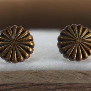 Op de afbeelding: Twee bronskleurige oorstekers in de vorm van een bloem. Elke oorbel heeft een gedetailleerd, geribbeld ontwerp dat uitstraalt vanuit een centraal punt. De oorbellen worden gepresenteerd op een wit oppervlak, met een houten rand eronder.