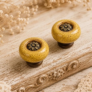 Puede incluir: Dos pomos de cerámica amarilla con acabado moteado y diseño floral en bronce oscuro en el centro. Los pomos están sobre una superficie de madera clara envejecida con tallas decorativas.