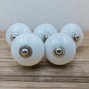 Peut inclure: Cinq boutons de meuble en céramique blanche avec une finition brillante. Chaque bouton présente un centre décoratif de couleur argentée. Les boutons sont disposés sur une surface en bois.