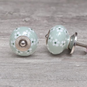 May include: Two light green ceramic cabinet knobs with white and green floral patterns. Each knob has a silver-colored base and screw. The knobs are round and have a smooth, glossy finish. Decorative hardware for furniture.