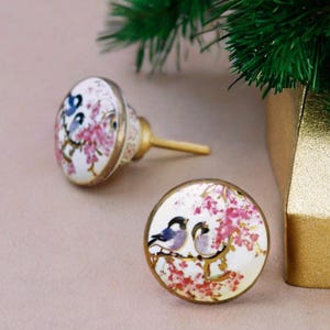 Op de afbeelding: Twee ronde keramische knoppen voor lades met een gouden metalen basis. De knoppen hebben een witte achtergrond met roze bloemen en blauwe vogels.