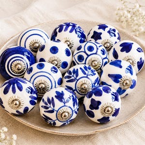 Puede incluir: Un conjunto de pomos de cerámica para armarios con diseños azules y blancos, incluyendo lunares, espirales y motivos florales. Cada pomo tiene una base de metal plateado. Los pomos están dispuestos sobre un plato de color neutro.