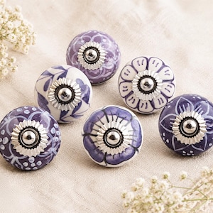 May include: Six decorative ceramic cabinet knobs with floral designs in shades of purple and white. Each knob has a silver metal base. The knobs are arranged on a light-coloured fabric. The knobs are suitable for cabinets.