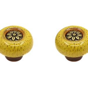 Könnte beinhalten: Zwei gelbe Keramik-Schrankknöpfe mit gesprenkelter Oberfläche. Jeder Knopf hat ein bronzefarbenes Blumendesign in der Mitte und einen dunkelbraunen Sockel. Diese dekorativen Knöpfe sind ideal, um Möbeln einen Hauch von Stil zu verleihen.
